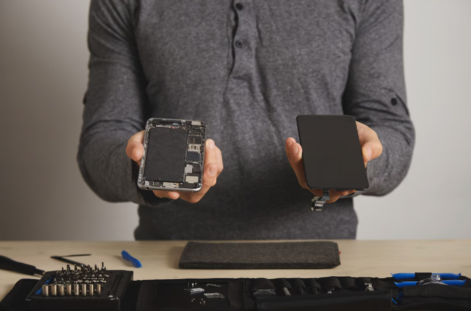 Technician holding a refurbished smartphone and replacement screen with repair tools on a desk