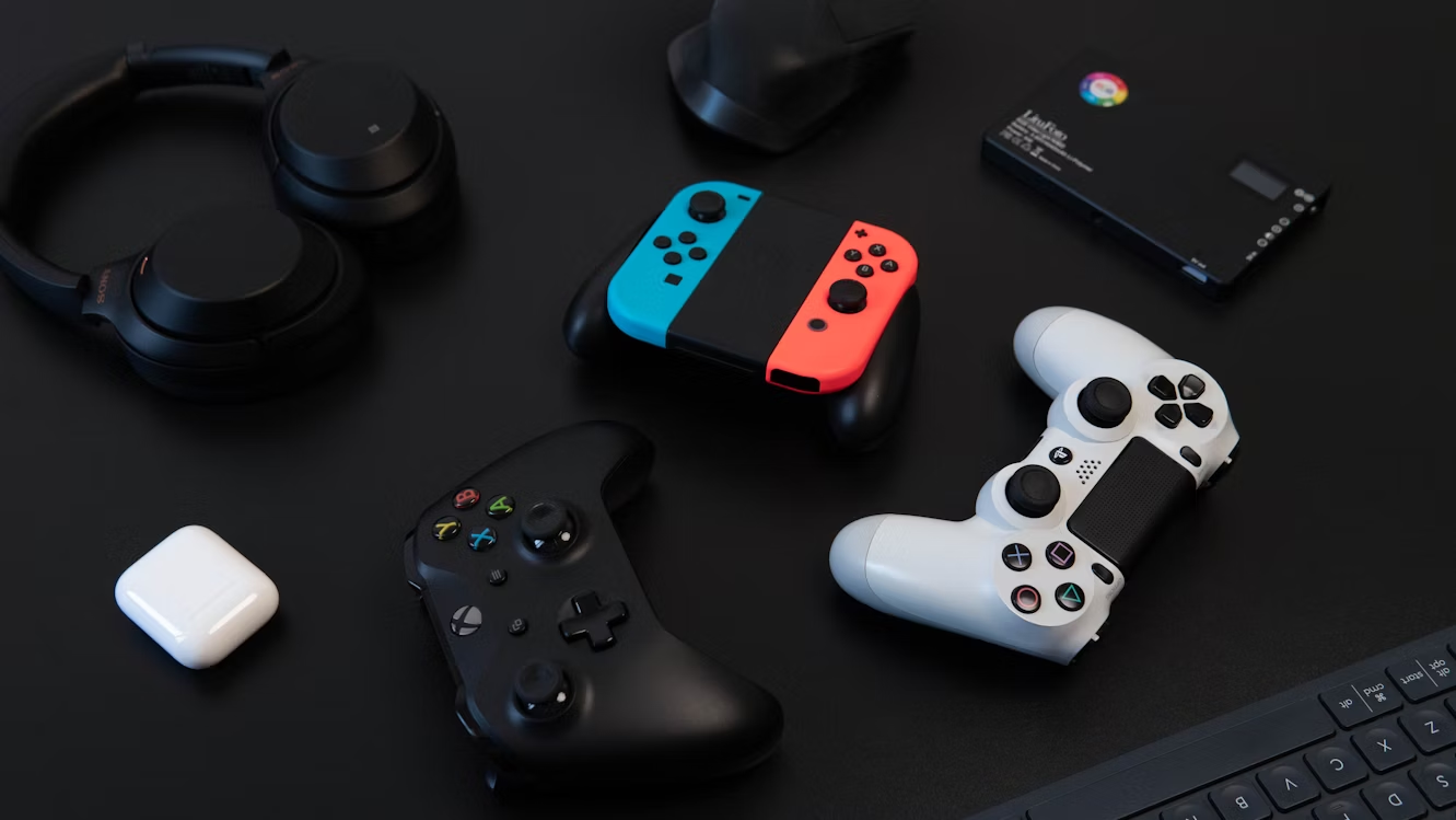 Flat lay of gaming accessories including Xbox controller, PlayStation controller, Nintendo Switch, headphones, and AirPods on black desk.